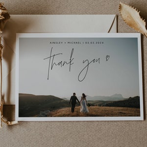 Pu&ograve; includere: Un biglietto di ringraziamento per un matrimonio con una foto di una coppia che si allontana dalla telecamera su una collina. Il biglietto dice "Ainsley & Michael | 03.02.2024" e "Grazie" in corsivo.
