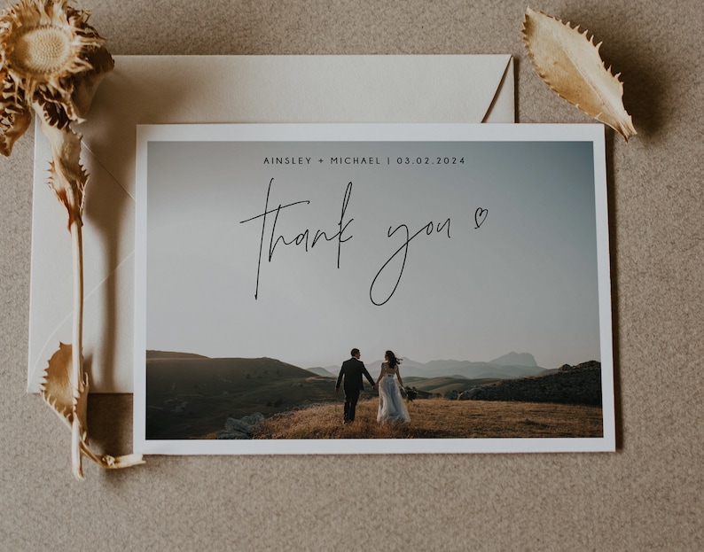 Pu&ograve; includere: Un biglietto di ringraziamento per un matrimonio con una foto di una coppia che si allontana dalla telecamera su una collina. Il biglietto dice "Ainsley & Michael | 03.02.2024" e "Grazie" in corsivo.