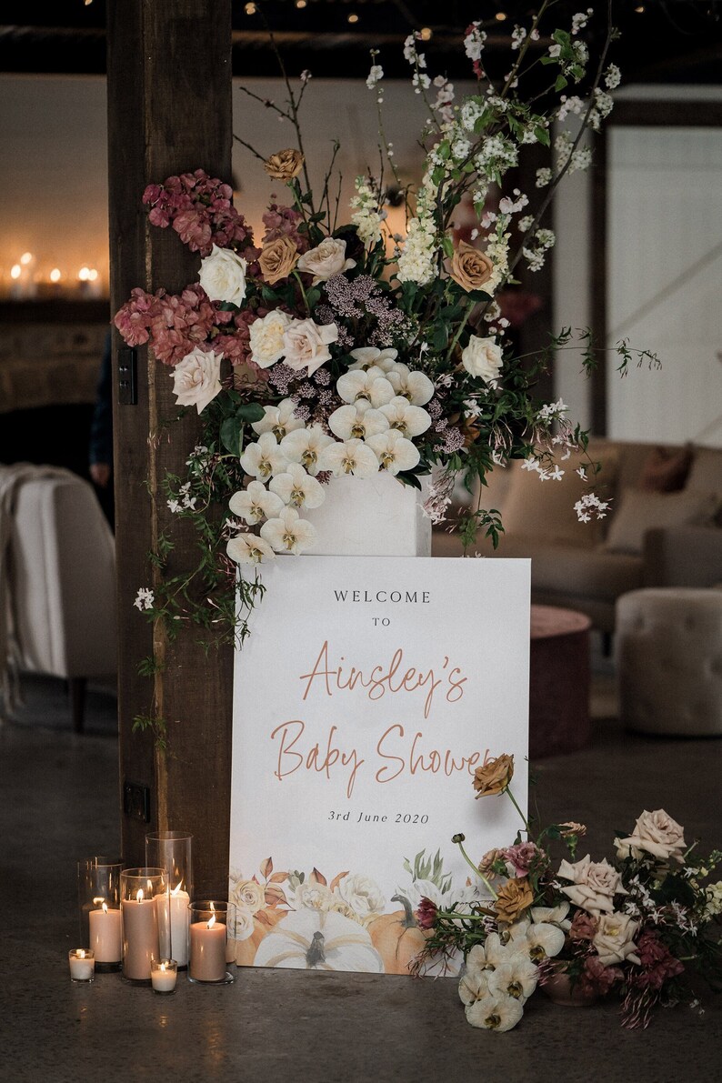 Op de afbeelding: Een welkomstbord voor een babyshower met de tekst "Welcome to Ainsley's Baby Shower 3rd June 2020". Het bord is versierd met aquarel pompoenen en bloemen. Het bord is omringd door kaarsen en bloemen.