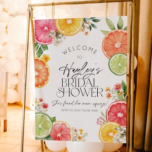 May include: A white bridal shower sign with a citrus theme. The sign says "Welcome to Hayley's Bridal Shower. She's found her main squeeze! We're so glad you're here!"