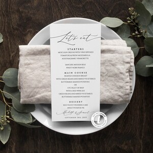May include: A wedding menu card with the words "Let's eat" in elegant script. The menu lists starters, main courses, and dessert options. The card is on a white plate with a linen napkin, with eucalyptus leaves in the background.