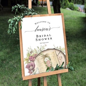 May include: A white sign with a floral design and an open book with a gold ring on a wooden background. The sign says "Welcome to Lauren's Bridal Shower 18th August 20xx".