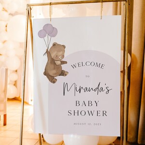 Puede incluir: Un cartel de bienvenida para un baby shower con un oso de dibujos animados sosteniendo globos morados. El cartel dice "Bienvenido a Miranda's Baby Shower 12 de agosto de 2023".