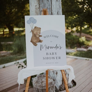 Puede incluir: Bienvenido a la Baby Shower de Miranda cartel con un oso marr&oacute;n sosteniendo globos azules. El cartel est&aacute; en un caballete de madera y tiene un fondo blanco con texto negro. La fecha de la ducha es el 12 de agosto de 2023.