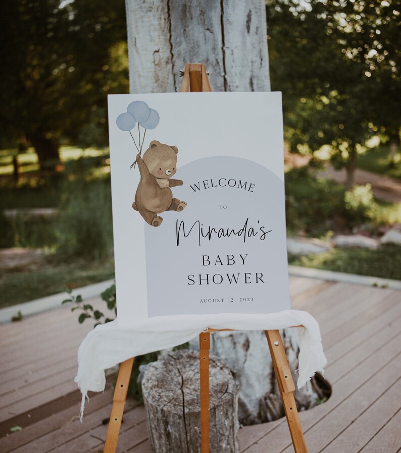 Puede incluir: Bienvenido a la Baby Shower de Miranda cartel con un oso marr&oacute;n sosteniendo globos azules. El cartel est&aacute; en un caballete de madera y tiene un fondo blanco con texto negro. La fecha de la ducha es el 12 de agosto de 2023.