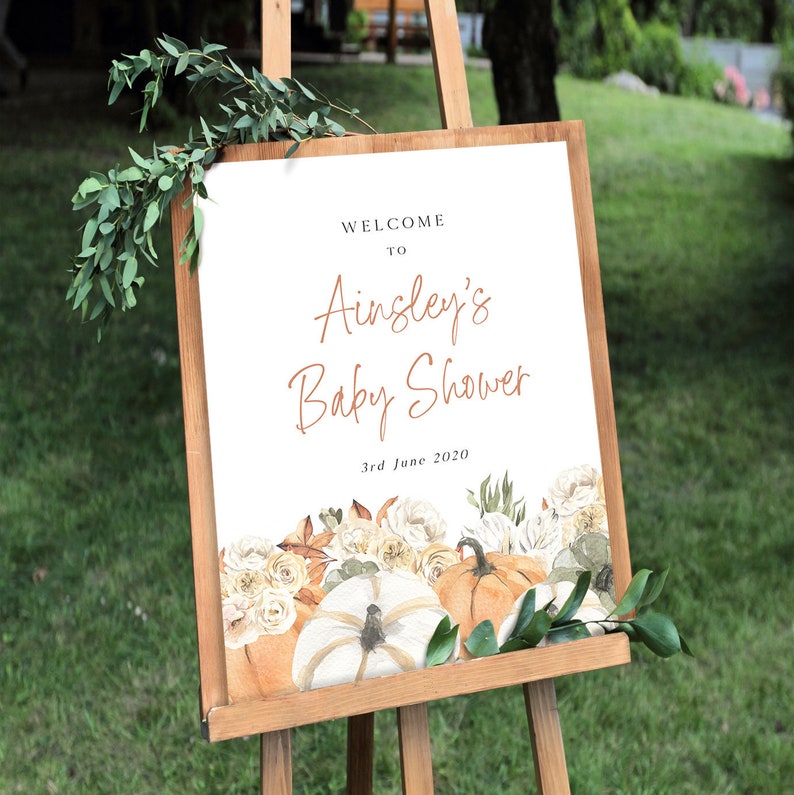 Op de afbeelding: Welkom bij Ainsley's Baby Shower bord met een aquarelontwerp van pompoenen en bloemen. Het bord staat op een houten ezel en de datum is 3 juni 2020.