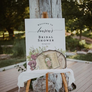 May include: A white sign with the text "Welcome to Lauren's Bridal Shower 18th August 20xx" and a watercolor illustration of an open book with flowers and a ring on a tree stump.