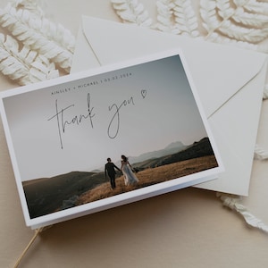 Pu&ograve; includere: Un biglietto di ringraziamento bianco con una foto di una coppia che cammina in lontananza sulla cima di una collina. Il testo sul biglietto recita "Ainsley + Michael | 03.02.2024" e "grazie" con un cuore.