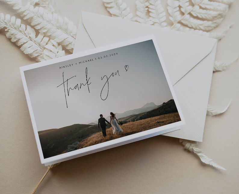 Pu&ograve; includere: Un biglietto di ringraziamento bianco con una foto di una coppia che cammina in lontananza sulla cima di una collina. Il testo sul biglietto recita "Ainsley + Michael | 03.02.2024" e "grazie" con un cuore.