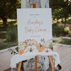 Op de afbeelding: Welkom bij Ainsley's Baby Shower bord met een aquarelontwerp van pompoenen en bloemen. Het bord staat op een houten ezel en de datum is 3 juni 2020.