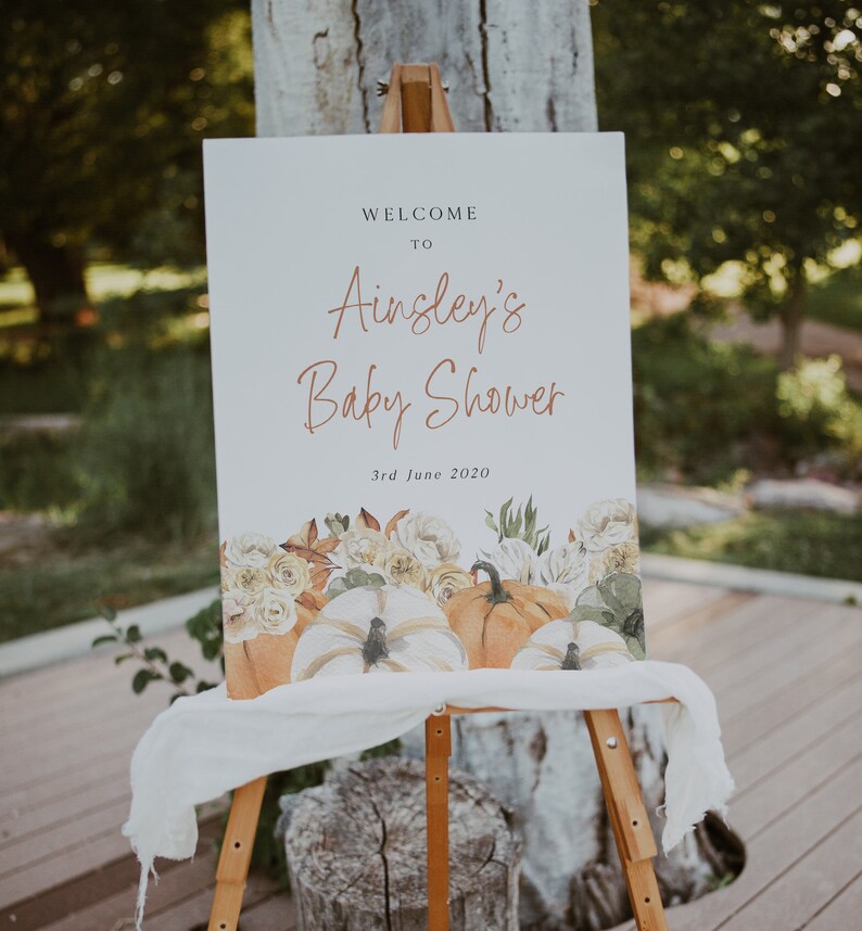 Op de afbeelding: Welkom bij Ainsley's Baby Shower bord met een aquarelontwerp van pompoenen en bloemen. Het bord staat op een houten ezel en de datum is 3 juni 2020.