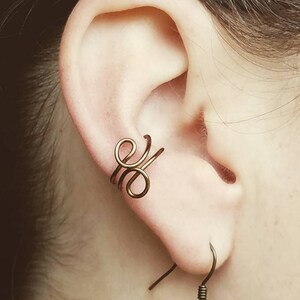 May include: A close-up of a person's ear with a gold ear cuff and a gold dangle earring. The ear cuff is a simple design with a swirl pattern. The dangle earring is a long, thin, cone-shaped pendant.