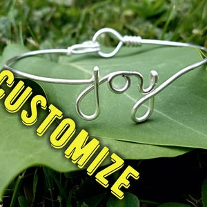 May include: Silver wire bracelet with the word "joy" formed into the design. The bracelet is on a green leaf with the word "CUSTOMIZE" in yellow letters.