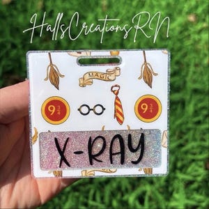 May include: A square badge reel featuring a white background with a pattern of broomsticks, magic scrolls, round glasses, striped ties, and platform 9 3/4 signs. A rectangular insert at the bottom displays the black text X-RAY on a glittery background.