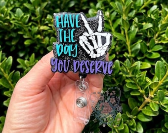 Może przedstawiać: Czarny, brokatowy identyfikator z napisem "Have the day you deserve" w kolorze turkusowym i fioletowym. Projekt zawiera szkielet dłoni wykonujący znak pokoju. Identyfikator ma przezroczysty plastikowy uchwyt i srebrny guzik.