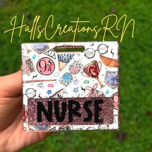 May include: A white badge reel with a glittery pink border and the word "NURSE" in black. The badge features colorful illustrations of Harry Potter-themed elements. The text "Halls Creations RN" is written in yellow script.