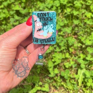May include: A rectangular badge reel with a glittery teal background and the text "DON'T TOUCH ME! I'M STERILE!" features a cartoon Patrick Star character wearing a doctor's head mirror and blue gloves. The reel has a clear plastic clip.