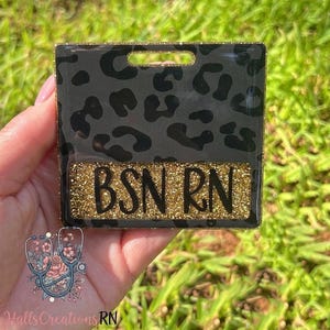 May include: A square badge holder featuring a black and dark gray leopard print pattern with a gold glitter border. A rectangular gold glitter insert at the bottom displays the black text BSN RN. The badge holder has a slot at the top.