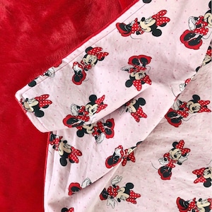 May include: A red and pink fleece blanket with a Minnie Mouse print. The blanket is reversible, with a solid red side and a pink side with white polka dots and Minnie Mouse images.