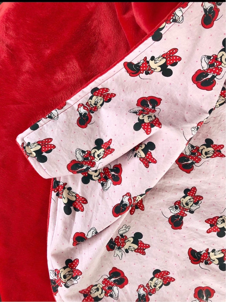 May include: A red and pink fleece blanket with a Minnie Mouse print. The blanket is reversible, with a solid red side and a pink side with white polka dots and Minnie Mouse images.
