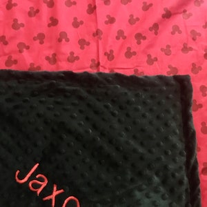 May include: Black and red personalized baby blanket with the name Jaxon embroidered in red on the black side. The other side of the blanket is red with a repeating pattern of black Mickey Mouse heads.
