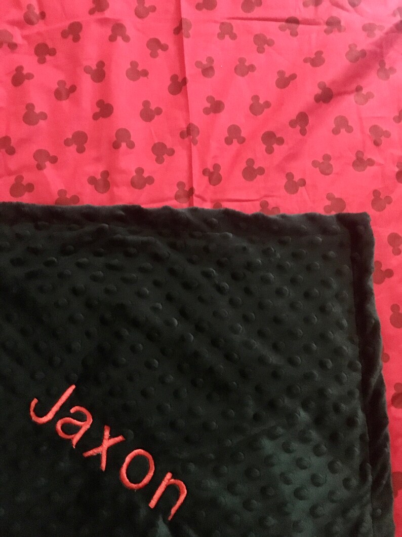 May include: Black and red personalized baby blanket with the name Jaxon embroidered in red on the black side. The other side of the blanket is red with a repeating pattern of black Mickey Mouse heads.
