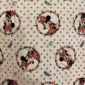 May include: White fabric with a repeating pattern of Minnie Mouse in a red dress with white polka dots. Each Minnie Mouse is surrounded by a wreath of flowers and leaves. The background is covered in small black dots.