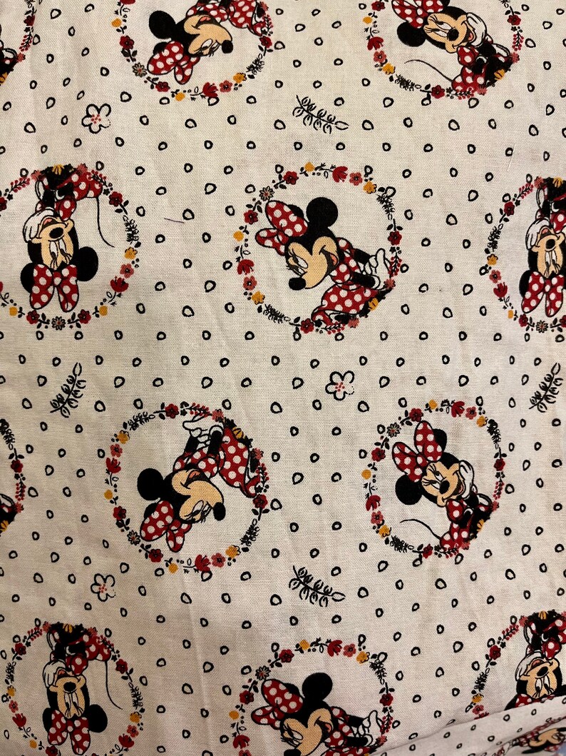 May include: White fabric with a repeating pattern of Minnie Mouse in a red dress with white polka dots. Each Minnie Mouse is surrounded by a wreath of flowers and leaves. The background is covered in small black dots.