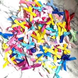 May include: A large assortment of colorful satin ribbon bows in various shades of blue, pink, yellow, red, and white. The bows are arranged on a white marble surface.