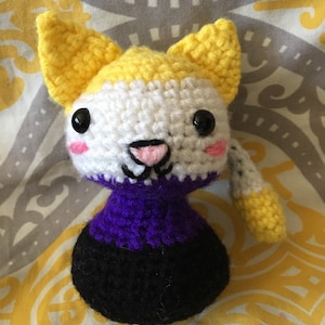 May include: A crocheted plush toy cat with yellow, white, purple, and black yarn. The cat has a pink nose and black eyes.