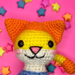 May include: A crocheted cat toy with orange, yellow, and white fur. The cat has black eyes and a pink nose. The cat is wearing a blue and white striped shirt. The cat is surrounded by colorful origami stars.