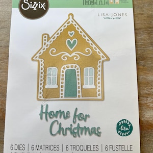 May include: A Sizzix Thinlits die set featuring a gingerbread house design. The house is gold with white trim, a teal door and heart accents. The words "Home for Christmas" are in teal script. The package includes 6 dies.