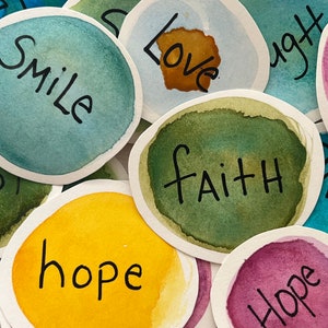 May include: A set of colorful, round, watercolor painted paper circles with inspirational words written in black ink. The words include "let go", "smile", "love", "faith", "hope", and "light".