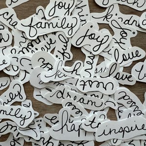 May include: A collection of white paper stickers with black handwritten words on them. The words include "create", "change", "joy", "family", "faith", "kindness", "love", "dream", "happy", and "inspire".