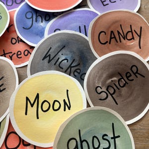 May include: A collection of watercolor painted circles with Halloween-themed words in black script. The circles are in various colors, including orange, purple, brown, and yellow. Words include "Pumpkin," "Eek," "Candy," and "Spider."