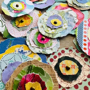 May include: A collection of layered paper flowers in various colors and patterns, each featuring a central metallic brass-toned brad. The circular paper cutouts display diverse textures, including floral, geometric, and polka dot prints.