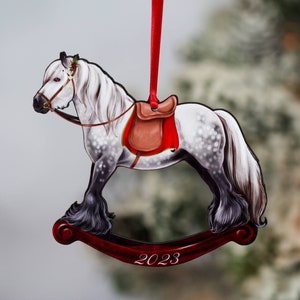 May include: A white and gray rocking horse Christmas ornament with a red saddle and a red ribbon. The ornament is on a red rocking base with the year 2023 printed on it.