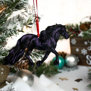 以下が含まれることがあります： 雪が積もった松の木に赤いリボンがついた黒い馬のクリスマスオーナメント。オーナメントは木でできており、光沢のある仕上げになっています。
