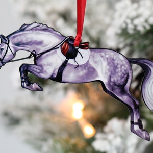 May include: A purple and white horse ornament with a red saddle and bridle, jumping over an obstacle. The ornament is made of metal and has a shiny finish.