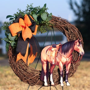 Pu&ograve; includere: Una ghirlanda di rami marroni con un fiocco nero e arancione e un ritaglio di un cavallo marrone e bianco con una sella. Il cavallo &egrave; in piedi su un supporto di metallo verde.