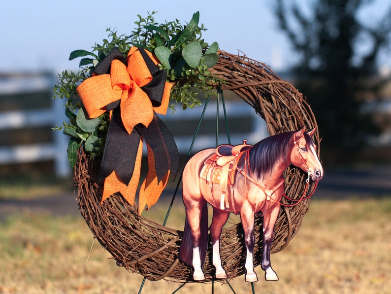 Pu&ograve; includere: Una ghirlanda di rami marroni con un fiocco nero e arancione e un ritaglio di un cavallo marrone e bianco con una sella. Il cavallo &egrave; in piedi su un supporto di metallo verde.