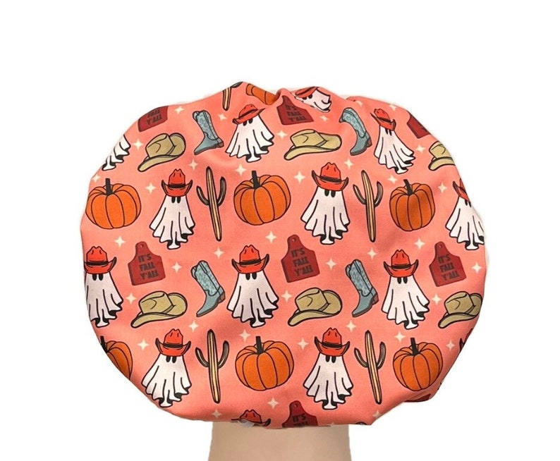 May include: A round shower cap with a peach-colored background featuring a repeating pattern of white ghosts wearing orange cowboy hats, orange pumpkins, blue cowboy boots, brown cacti, and red tags with the text IT'S FALL Y'ALL and small stars.