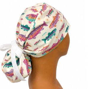 May include: A scrub cap with a cream-colored background featuring a repeating pattern of colorful fish in shades of pink, purple, and teal, accented by small orange suns and green waves. The cap includes a white ribbon tie at the back for adjustment.