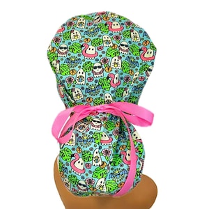 May include: A teal scrub cap with a fun ghost and flamingo print. The cap features a pink ribbon tie and is designed to be worn on the head. The fabric is patterned with cartoon ghosts wearing sunglasses and floating on inflatable flamingos.