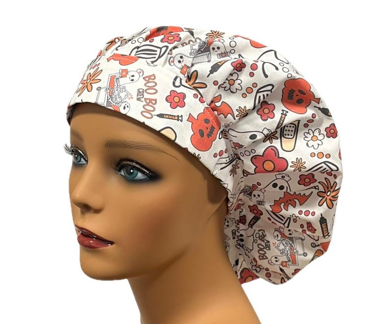 Op de afbeelding: Witte scrub-muts met een Halloween-print met geesten, pompoenen, vleermuizen en de tekst "Boo Boo Crew".