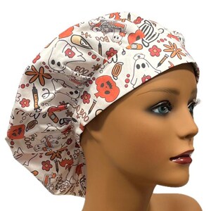 Op de afbeelding: Witte scrub-muts met een Halloween-print met geesten, pompoenen, spuiten en de woorden "Boo" en "Boo Boo".