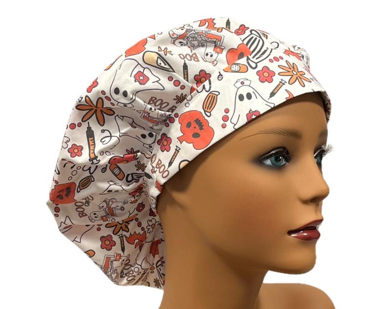 Op de afbeelding: Witte scrub-muts met een Halloween-print met geesten, pompoenen, spuiten en de woorden "Boo" en "Boo Boo".