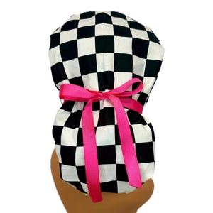 May include: A black and white checkered scrub cap with a bright pink ribbon tied in a bow. The cap is designed to be worn on the head and has a gathered back. The pink ribbon adds a pop of color to the design.
