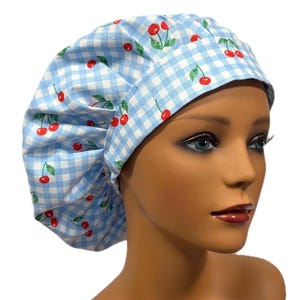 May include: A light blue and white gingham patterned bouffant scrub cap. The cap is decorated with red cherries and green leaves. The cap is worn by a mannequin with a fair complexion and red lipstick.