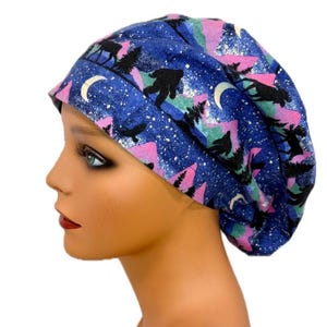 May include: A scrub cap featuring a dark blue background with a repeating pattern of black silhouettes of Bigfoot, moose, and pine trees, accented by pink and teal mountain shapes, white crescent moons, and a subtle silver glitter effect throughout.